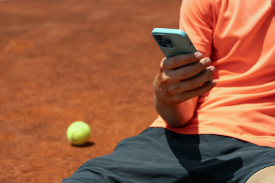 Close-up of young tennis player using smartphone during break on clay court with tennis ball nearby and copy space for sports technology advertising