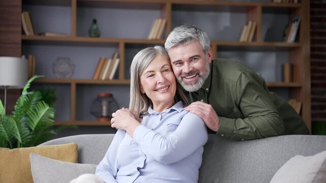 Senior husband gently hugging his wife while she sits on a sofa in the living room at home. Smiling retired couple sharing kind words, affection and emotional support during a warm, intimate moment.