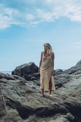 Woman in a long beige dress stands alone on dark rocks looking out toward the open sea. Contemplation and solitude by the sea supporting mindfulness and slow travel themes © Екатерина Лукашина
