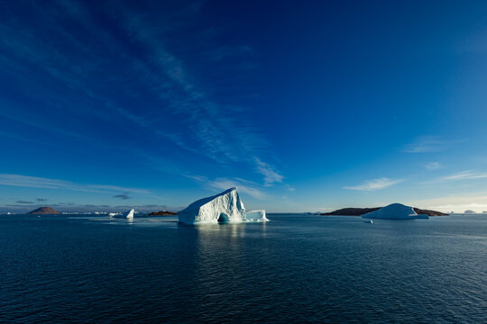 Bright blue iceberg reflects in calm water with cloudy sky and distant mountains along the coast of Greenland