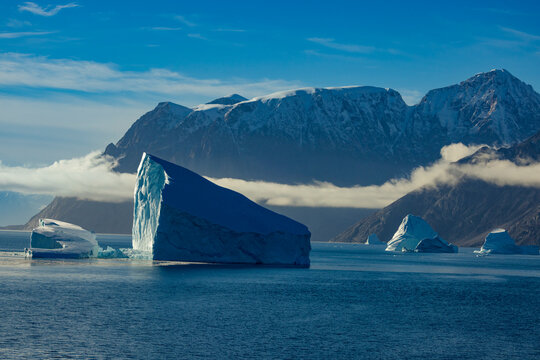 Towering icebergs rise against mountain peaks in golden Arctic light with clouds in Scoresby Sund East Greenland