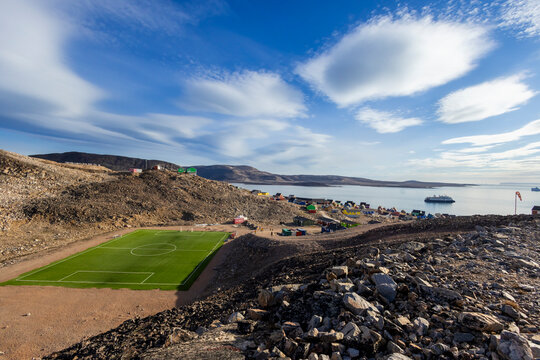 View of Ittoqqortoormiit settlement with soccer field surrounded by rocky terrain and Arctic Ocean