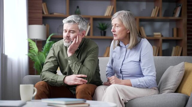 Senior wife confronting and nagging her husband while sitting on a sofa in living room at home. Retired man appears emotionally drained as couple experiences ongoing relationship conflict and tension.
