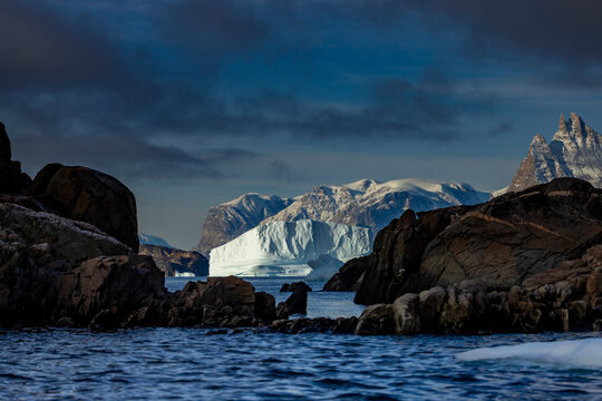 Icy waves crash on rocky shores with a towering iceberg in the distance near dark clouds and mountain peaks