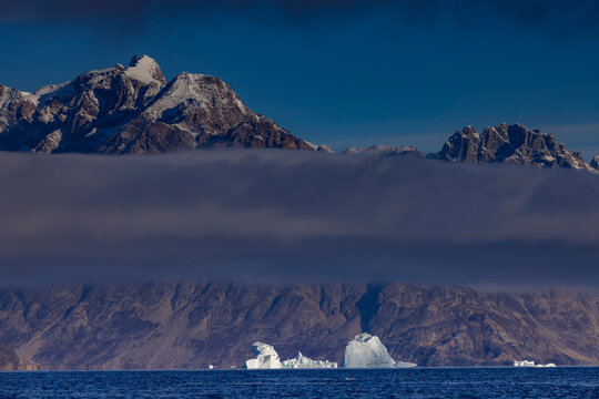 Icebergs float in the ocean beneath a striking cloud band over Warming Island in Greenland