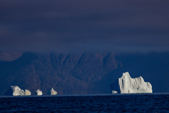 Icebergs glow under dark clouds with rocky island backdrop in Arctic waters of Greenland