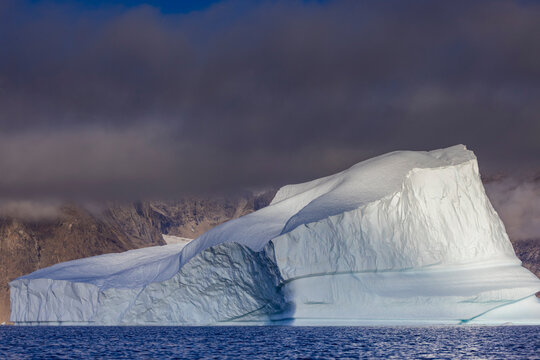 Monumental iceberg rises from the sea near rugged mountains under cloudy skies in Greenland