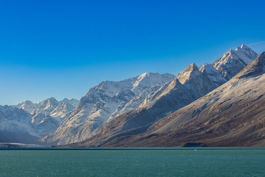 Snow-capped peaks rise over barren land and calm waters in the Arctic landscape of Blomsterbugten, Greenland under a clear sky