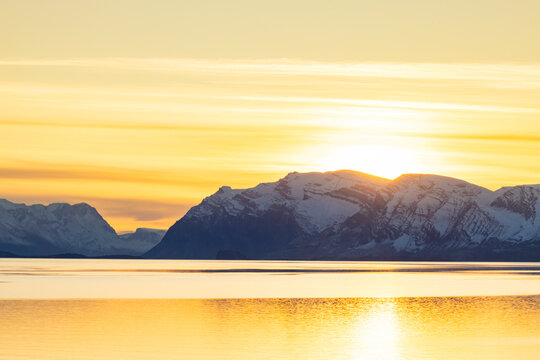 Golden light spreads over snow-covered peaks and a calm fjord during dusk in Greenland with the sun setting behind mountains