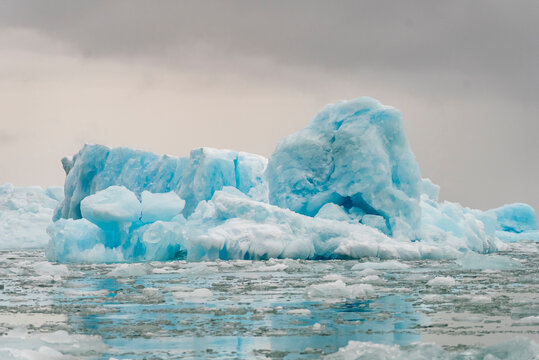 Sculpted iceberg rises from the frigid sea under overcast sky with soft curves and pale blue tones near broken drift ice in Greenland