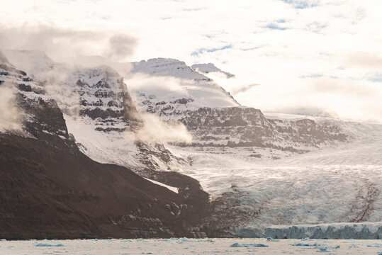 Mist drifts over snowfields and rocks as a glacier flows toward water in the Arctic light of Borgvig in Greenland