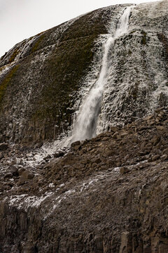 Narrow waterfall cascades down a frost-lined cliff in winter landscape with rocky terrain and snow