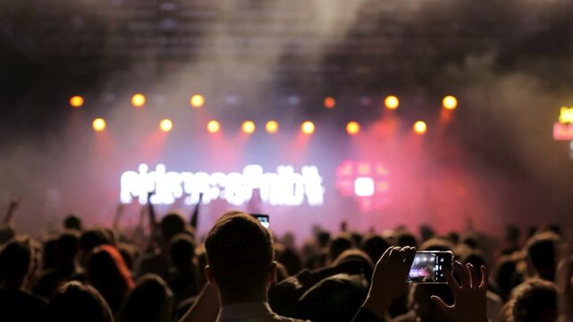 Fan records the band leaving a bright rock stage on a smartphone while the crowd applauds, capturing mobile memories and audience participation at the festival.
