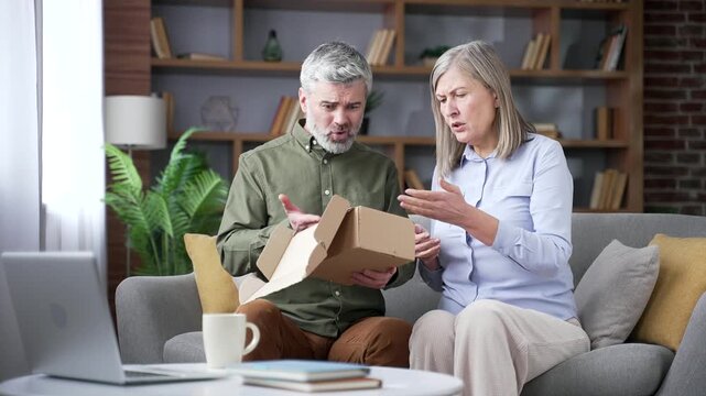 Disappointed senior married couple opening a delivery box while sitting on a sofa in living room at home. Retired husband and wife upset after receiving a wrong or damaged product from online order.