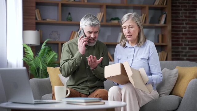Disappointed senior husband complaining on a smartphone after receiving a wrong product while sitting on a sofa in the living room at home. Retired couple dealing with poor online shopping service.