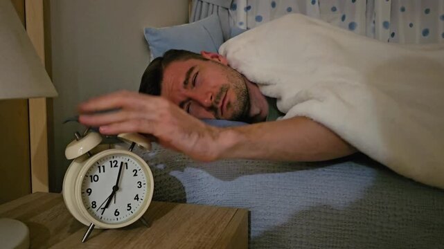 Adult man sleeping in bed, waking up to ringing alarm clock, reaching to switch off alarm and falling asleep again with hand resting on clock. Oversleeping, morning routine and sleep deprivation