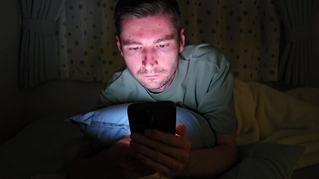 Adult man using smartphone while lying in bed in dark bedroom illuminated by mobile phone screen. Late night screen time, doomscrolling, insomnia, digital technology addiction and sleep disorder