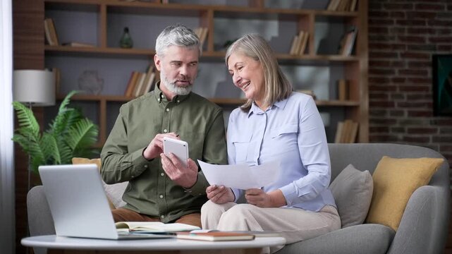 Senior married couple calculating household finances with a calculator while holding utility bills sitting in home office. Happy retired husband and wife smiling after discovering they had saved money