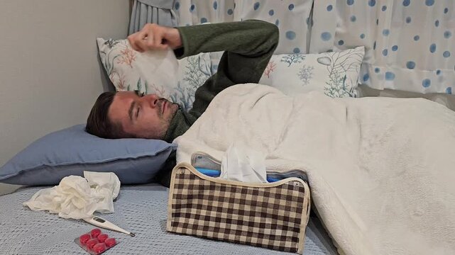 Sick man lying in bed with a cold compress on his forehead, surrounded by tissues, thermometer and medicine. Home illness recovery footage showing fever, flu symptoms and rest in bedroom