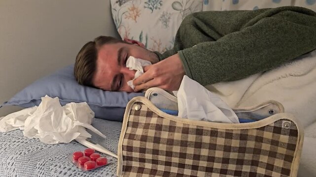 Sick young man lying in bed blowing nose with tissue beside medicine and tissue box. Home recovery footage showing cold or flu symptoms, seasonal illness and resting indoors