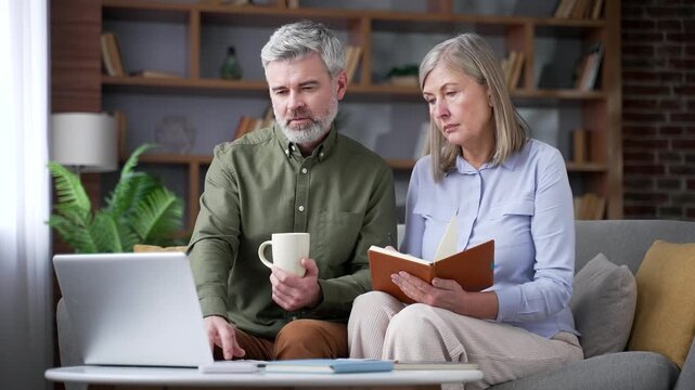 Senior married couple taking notes while watching an online webinar on a laptop in the living room at home. Retired husband and wife learning new skills through a virtual course and presentation.