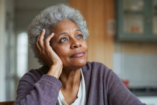 Soft light illuminates a thoughtful senior black woman. Her hand rests gently on her cheek. Glasses, purple cardigan, and gray curls frame her calm gaze. She looks upward, lost in quiet reflection