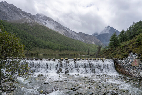 Water flows over a small dam in a high-altitude valley surrounded by mountains and forest in Yading Nature Reserve Sichuan China