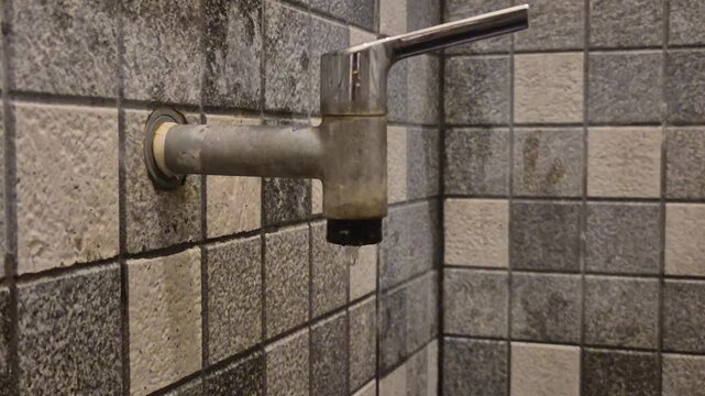 A close-up shot of an old, tarnished chrome water tap installed on a wall with grey and beige mosaic tiles in a bathroom or washroom, showing signs of water stains and limescale buildup.