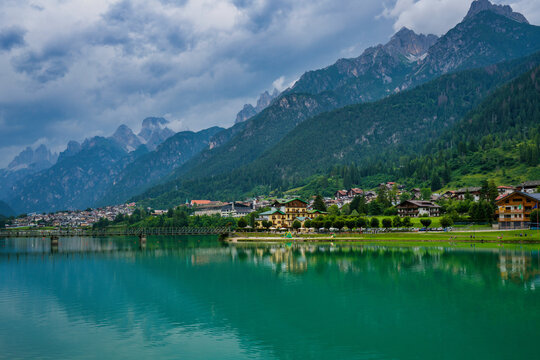 Mountains and village reflect on emerald waters in Auronzo, Veneto, Italy during cloudy weather