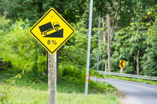 Steep Incline Warning Sign: A diamond-shaped yellow warning sign indicates a steep 8% incline for trucks on a winding road surrounded by lush green trees and vegetation.