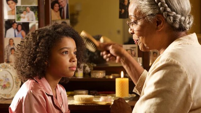 Reaching with wooden brush, grandma brushing girl's curls in pink satin pajamas at vanity for care