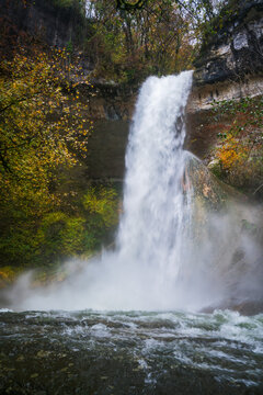 Sugarloaf waterfall flows over rocks surrounded by autumn leaves in Surjoux, Ain, France during the fall season