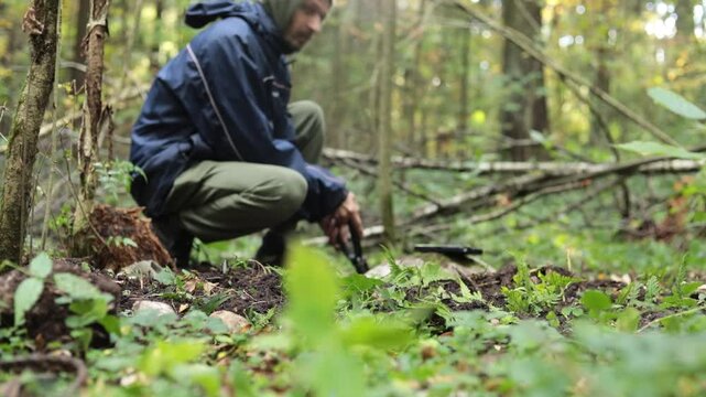 A guy digs up an object in the woods. A man digs a rock. A man makes a hiding place in the wilderness. An unknown man is looking for something in the ground.