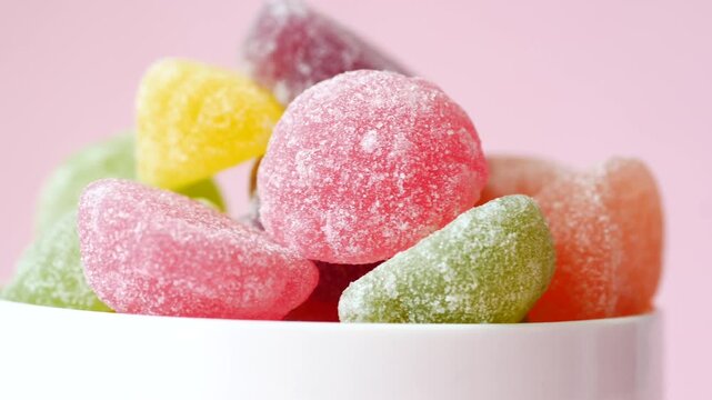 Close up rotation of colorful gummy candies in white bowl against pink background showcasing sugar coating and textures