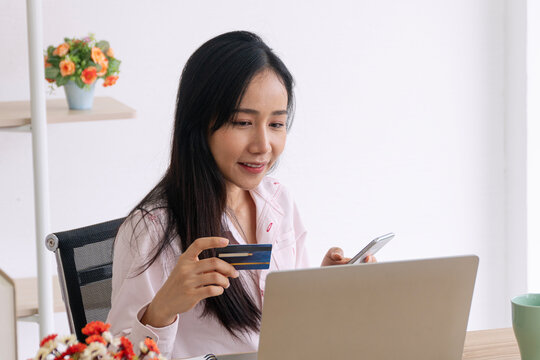 Smiling young Asian woman holding a credit card and using a smartphone at a desk with a laptop. Happy female consumer shopping online and making quick digital payments from a bright home office.