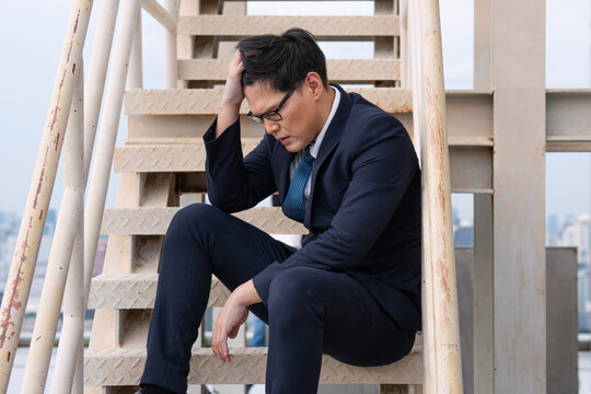 Stressed Asian businessman in a suit sitting on outdoor metal stairs holding his head. Depressed corporate executive feeling exhausted, overwhelmed, or facing a financial crisis with city background.