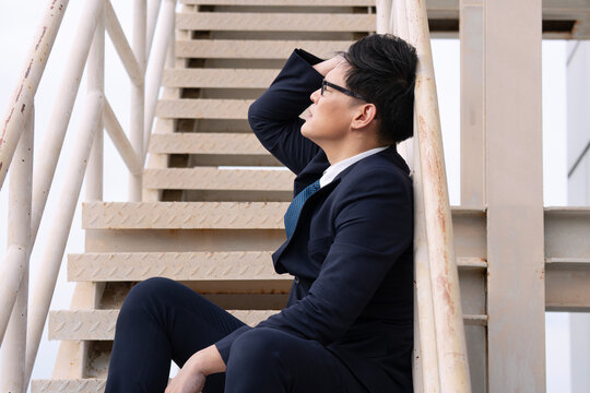 Stressed Asian businessman in a dark suit sitting on outdoor metal stairs leaning his head back. Exhausted corporate executive experiencing burnout, financial crisis, or job loss.
