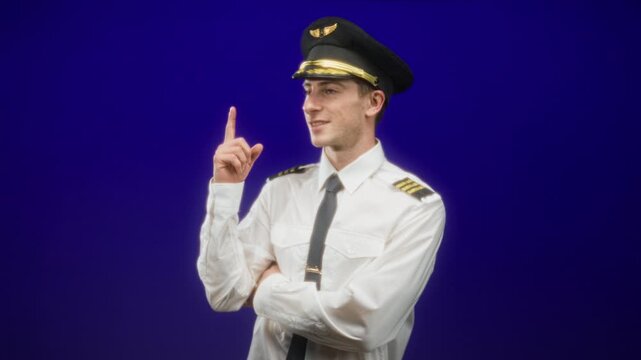 Man pilot in uniform points finger upward with crossed arms, showing cap and epaulettes in studio; confidence duty.