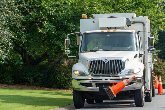 White International utility truck with an orange traffic cone secured to its front bumper, parked on a paved road with lush green trees and grass in the background on a bright day.