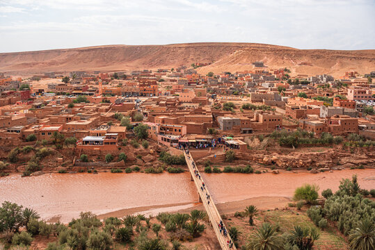 View of Ait Ben Haddou village with footbridge connecting old and new sections over reddish riverbanks in clear skies