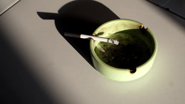 A close-up shot reveals a lit cigarette in a green ceramic ashtray on a white floor surface illuminated by sunlight, smoke curling upwards with ashes falling in time lapse video.