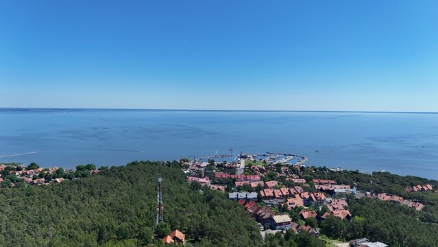 Aerial panoramic view of Nida village on the Curonian Spit, Lithuania. Scenic summer landscape of a coastal resort town with red-roofed houses, yacht harbor, green pine forest, and Curonian Lagoon.