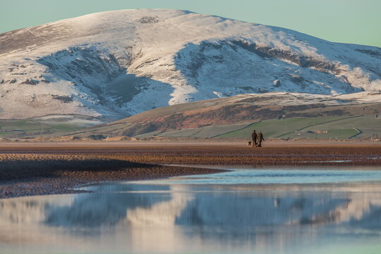 Snow covers Black Combe as people walk along the shoreline of Walney Island in Cumbria United Kingdom