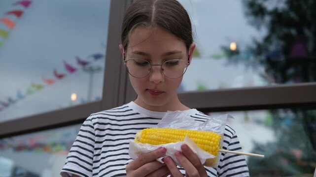 Young girl with glasses enjoys eating corn on the cob while standing outside a building decorated with colorful flags and festive elements in the background