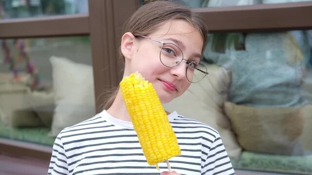 Young girl with glasses enjoys corn on the cob, transitioning from a thoughtful expression to a cheerful smile, seated outside a cafe with soft furnishings in the background