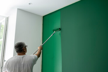 Professional painter applying deep green paint with roller on modern interior accent wall during home renovation in bright residential room with natural light, copy space. © JairoMZ