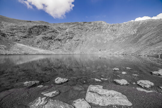Flat calm Scales Tarn reflects Sharp Edge under blue skies in the English Lake District, popular route to Blencathra summit