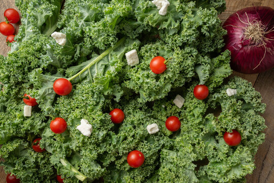 Fresh green kale, (Brassica oleracea), vine ripened, red cherry tomatoes, feta cheese, and a whole purple onion spread out on a wooden table.