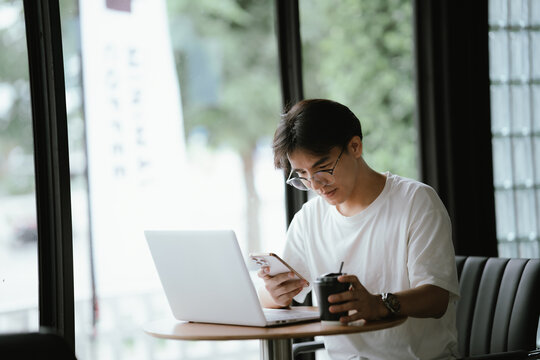 Young professional talking on smartphone while working on a laptop in a modern caf&eacute;, holding a coffee cup with natural window light.