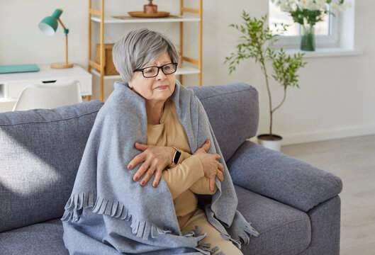 Portrait of upset senior woman sitting on sofa in living room at home wrapped in cozy blanket trying to keep warm. Elderly retired person freezing in winter in chilly room with low temperature.